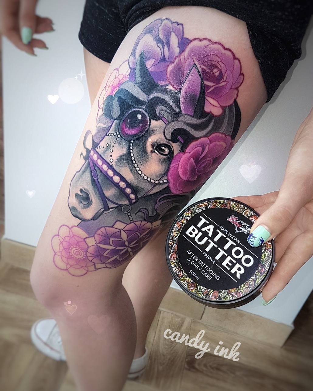 the image shows a woman's thigh with a colorful tattoo of a horse's head adorned with flowers and a unicorn horn.  the tattoo has purple, pink and gray tones. the tattoo artist is holding a tub of vegan tattoo butter for aftercare.  the background is blurred, suggesting the photo was taken in a tattoo parlor.