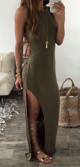a fullbody shot of a woman wearing an olive green maxi dress with side slits. the dress is sleeveless and simple. she is accessorized with gold gladiator sandals, a long gold necklace with a tassel pendant, and several bracelets. she carries a woven handbag.  the setting appears to be indoors with a beige couch and a tiled floor. the woman is posing for a photo, holding her phone.  her hair is long and brown. the overall style is bohochic.