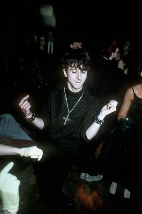 a young man with dark hair is dancing in a dark nightclub, wearing a black shirt and trousers. he is surrounded by other people, but they are out of focus. the atmosphere is lively and celebratory, with a sense of fun and excitement.