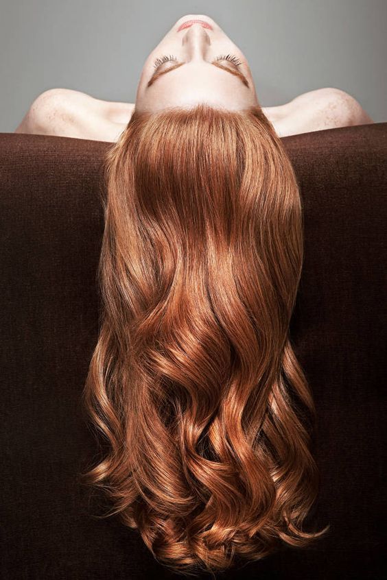 a beautiful woman with long, red, wavy hair poses for a portrait. the hair is styled in a loose, flowing style. the woman is lying on her back, her hair cascading down in front of her face. the background is a dark brown color, which creates a dramatic contrast with the woman's bright red hair. the woman's eyes are closed, and she appears to be in a state of relaxation and tranquility.