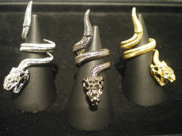 the image shows three rings in the shape of snakes. each ring is displayed on a black cone stand. from left to right, the rings are silver, gunmetal and gold. the snake's head is prominent on each ring. the background is a dark color.