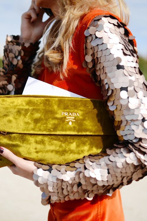 closeup of a woman holding a prada velvet clutch. she is wearing a bright orange dress with sequined sleeves. the image is taken outdoors in natural daylight. the overall style is autumnal/winter fashion.