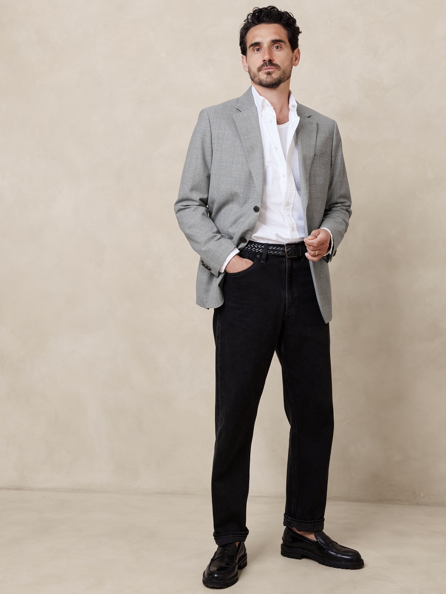 a full shot of a man standing with one hand in his pocket. he is wearing a gray blazer, a partially unbuttoned white shirt, black jeans, and black dress shoes. he has a black belt and dark hair.