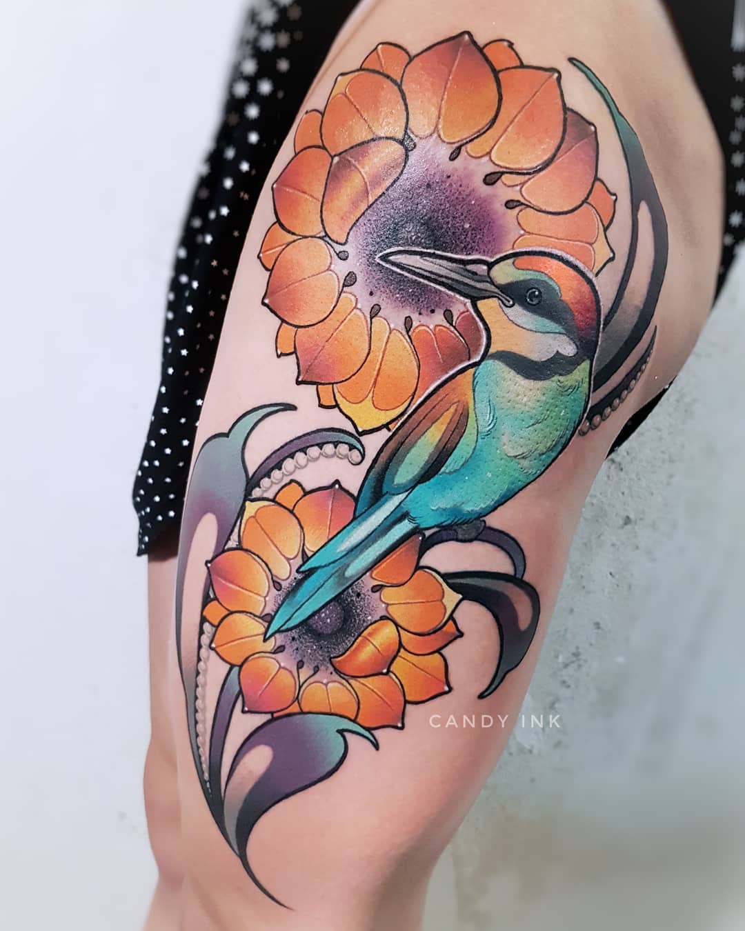 a person's upper left leg is shown with a colorful neo traditional tattoo of a blue and orange bird perched amongst orange flowers. the tattoo is on the outer thigh and is very detailed.