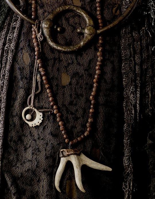 closeup shot of an assortment of earthy and rustic jewelry pieces against a dark, textured fabric backdrop. the jewelry includes a necklace with brown beads and an antler pendant, a metal ring, and a smaller bone pendant. the overall impression is one of antique or vintage items with a detailed and textured aesthetic.