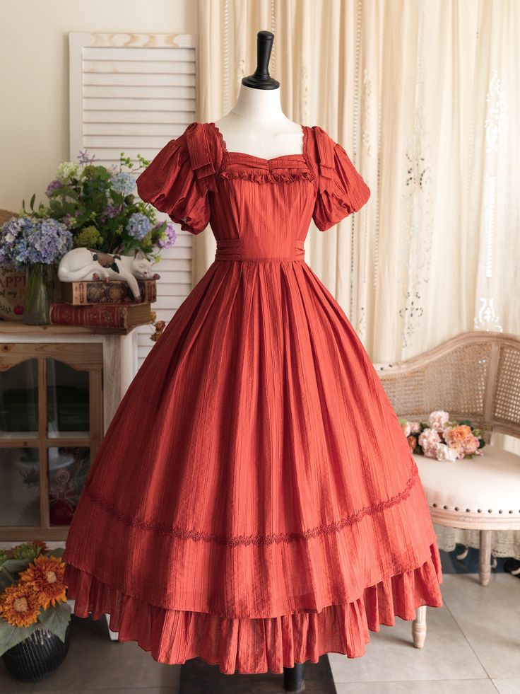 a fulllength image of a burnt orange vintagestyle dress displayed on a mannequin. the dress features a square neckline, puff sleeves, a cinched waist, and a flowing skirt with lace trim. the background includes a white wall, a window with sheer curtains, and a wooden cabinet adorned with flowers and books.  the lighting is soft and natural, highlighting the dress's intricate details.