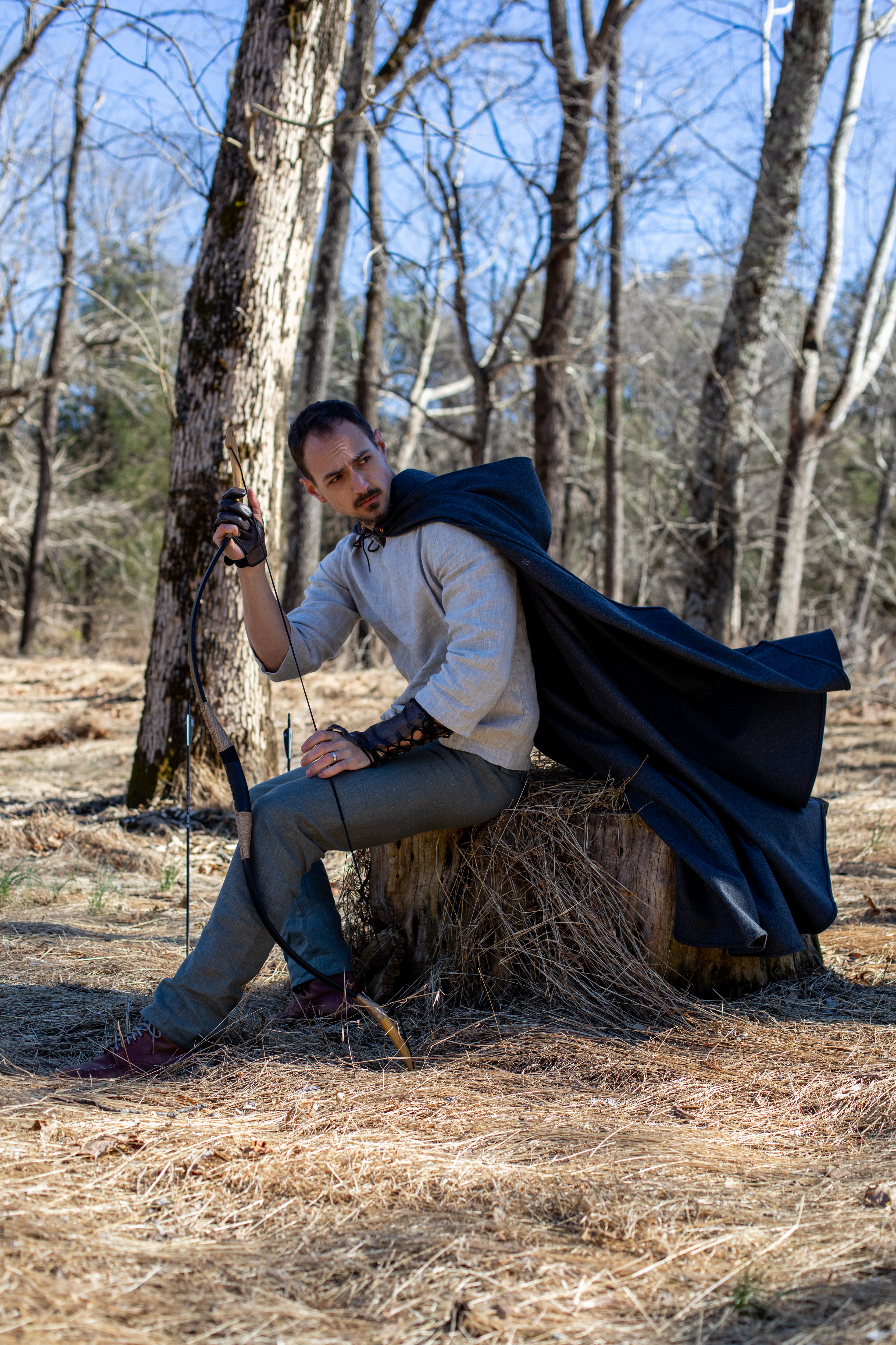 a man dressed in medieval fantasy attire, complete with a dark cloak and leather arm guards, sits perched on a tree stump in a sundrenched forest. he holds a bow in his left hand, arrows resting beside him, conveying an image of a ranger or hunter. the natural light filtering through the bare trees creates a sense of tranquility and adventure.