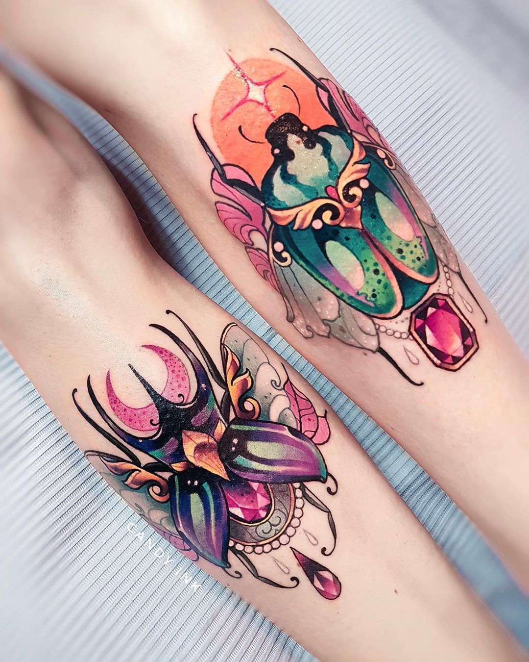 the image shows two neotraditional tattoos on someone's legs. the top tattoo depicts a green and gold jewel beetle with an orange sun behind it. the bottom tattoo shows a purple and gold scarab beetle with a crescent moon behind it. both beetles are adorned with jewels and crystals, and are surrounded by delicate linework. the overall color palette is warm and inviting, with pops of pink, teal, and purple.