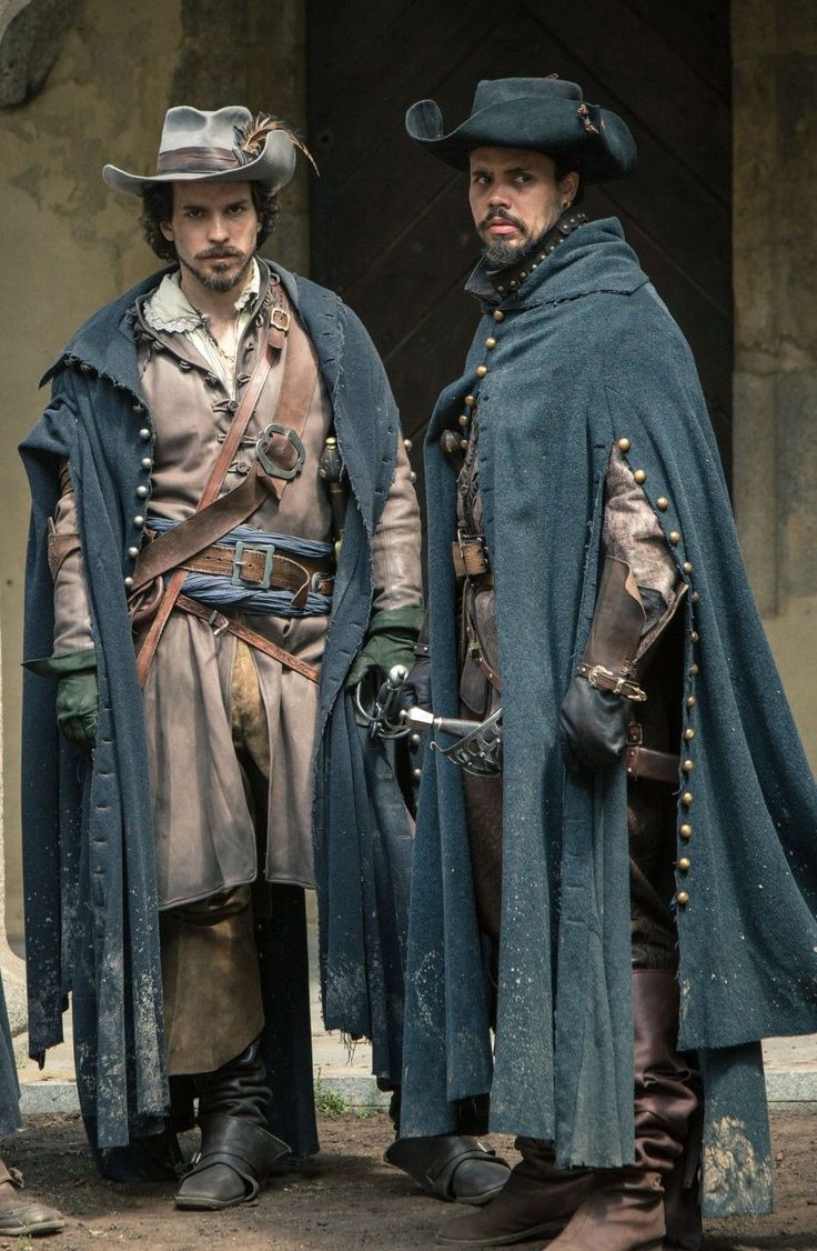 the image shows two men dressed in elaborate 17thcentury attire. they are wearing long, darkcolored cloaks, widebrimmed hats,  leather belts, and boots.  their clothing features buckles, belts, and other details characteristic of the period. they appear to be carrying swords or daggers. both men have serious expressions on their faces. the setting is outdoors, in front of a stone building. the overall style suggests a scene from a historical drama or a historical reenactment.  the costumes are detailed and authenticlooking.