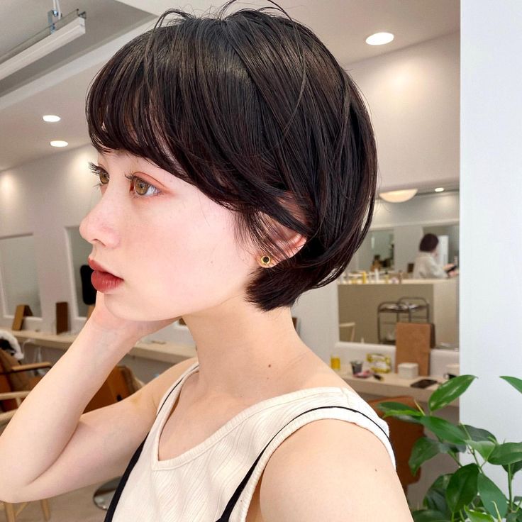 a close up side profile of a young woman with short black hair, styled in a bob with bangs. she is looking off to the side and has a slightly sad expression. the woman is wearing a white tank top and small golden earrings.