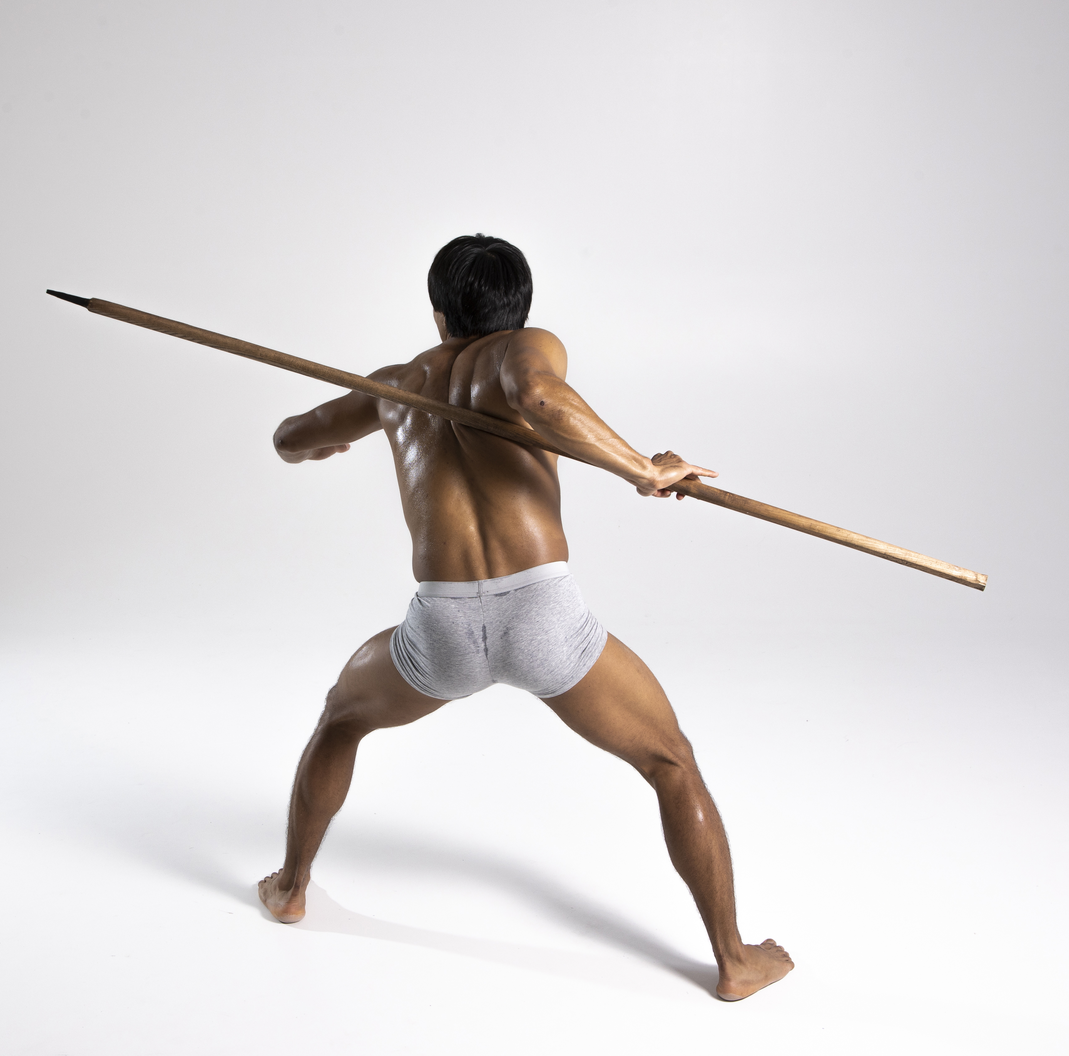 a muscular man is shown from the back in a studio shot against a white background. he's wearing grey briefs and is barefoot, holding a wooden spear horizontally across his back. the man is in a tense, readytostrike stance, highlighting his athletic build and strength.
