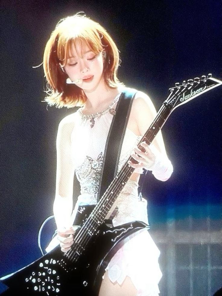 a young woman with short blonde hair is standing on stage in front of a microphone. she is playing a black electric guitar, and wearing a white, glittery, sleeveless top and a short white skirt. the background is dark, and she is illuminated by a bright spotlight.