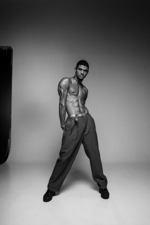 a black and white studio shot of a shirtless athletic man wearing pants and a belt. he is muscular and posing with his abs showing.