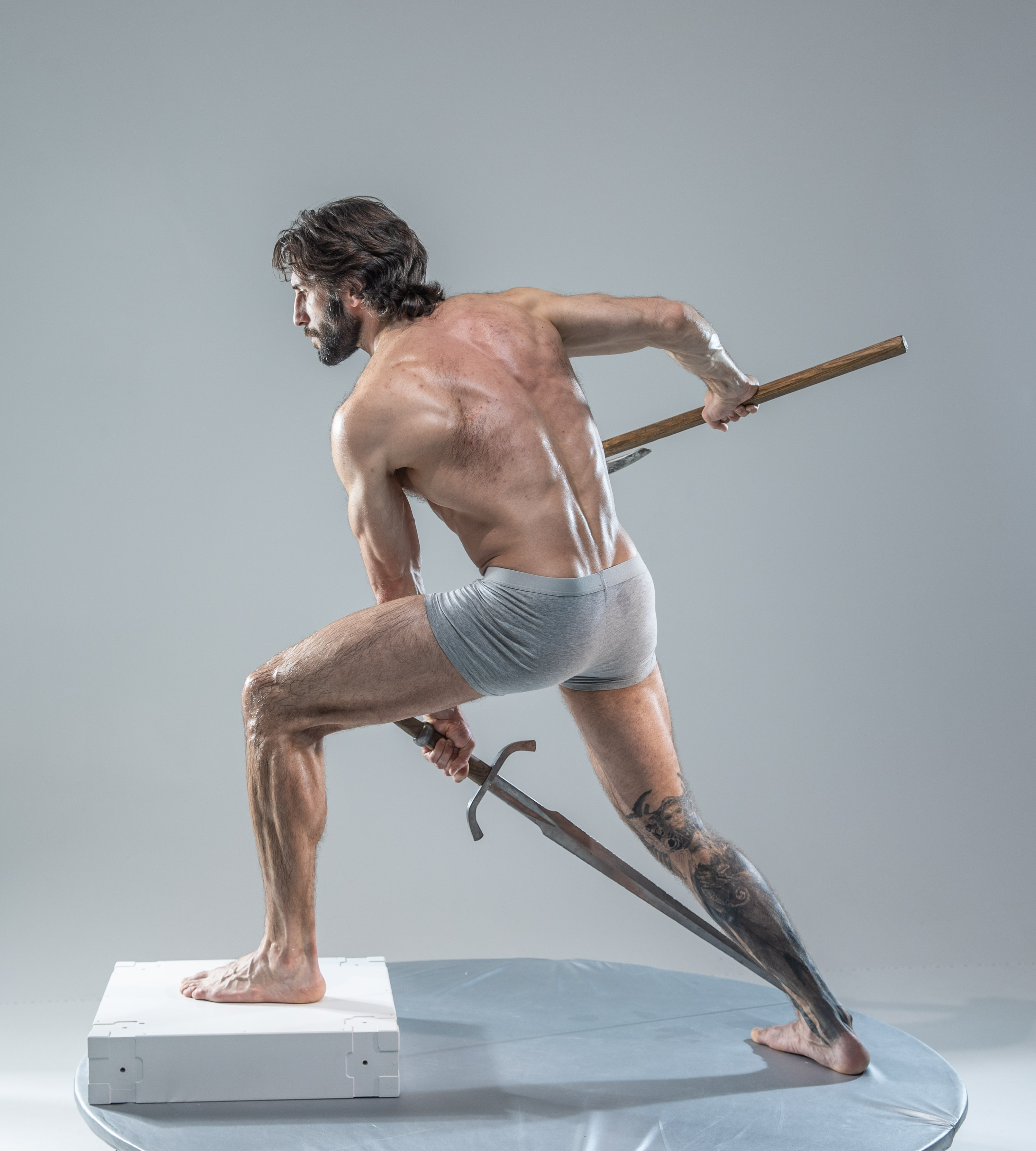 a muscular man with long hair and a beard poses on a white platform against a light grey background. he is wearing grey underwear and holds a sword in his right hand and a staff in his left, both made of wood. he is barefoot and his body is angled dynamically, as if he is about to strike or defend. the lighting is soft and diffused, highlighting his physique.