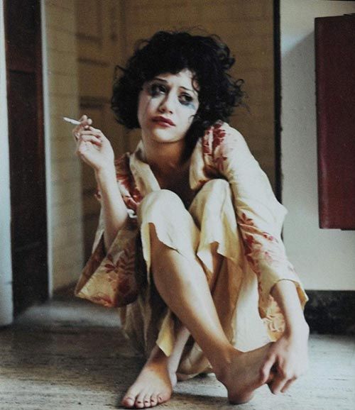 a young woman with dark hair sits on the floor with her legs crossed and her arms resting on her knees. she is looking at the camera and has a sad expression on her face. she is wearing a white robe with red flowers on it. the woman has black and white makeup on and is smoking a cigarette. the photo is in black and white.