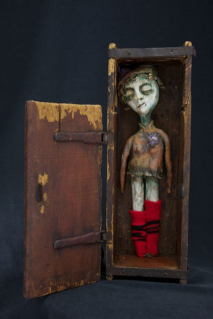 an antique, handmade doll stands inside an open wooden box with a latch. the doll has a slightly creepy and eerie appearance, with weathered features and wearing red socks with black stripes. the wooden box is old and vintagelooking, adding to the overall unsettling atmosphere of the image. the doll is standing up inside the box.