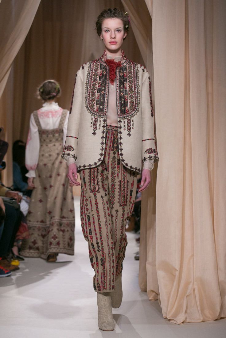 a woman model walks the runway in a white embroidered jacket and matching pants. she is wearing tan boots. the model's hair is pulled back and she has on light makeup. the background is out of focus, but you can see beige curtains and other models.