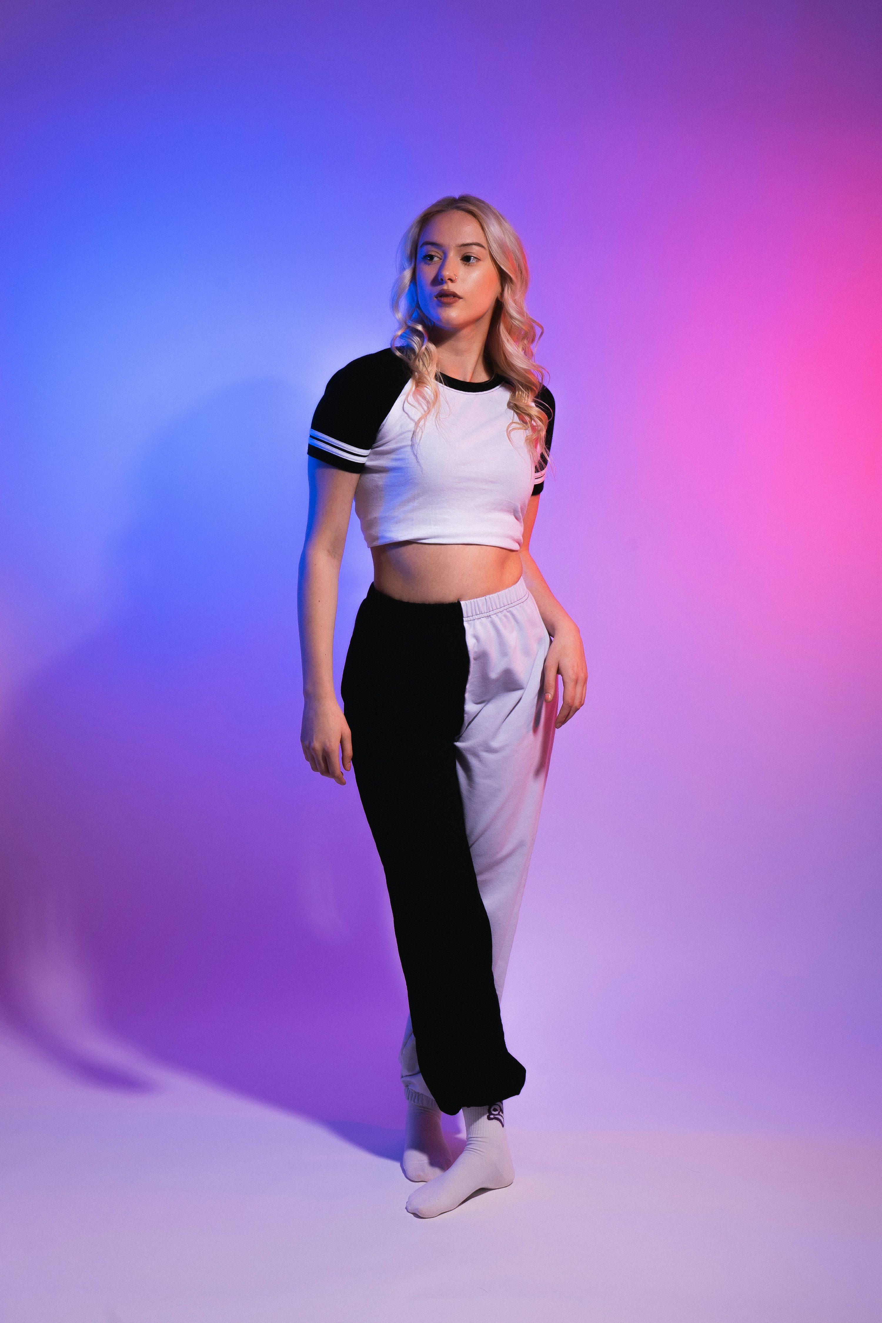 a fullbody shot of a young blonde woman posing in a studio against a vibrant purple and pink neon backdrop. she is wearing a white and black twotoned cropped top and matching sweatpants, white socks, and is styled in a casual yet trendy streetwear look.  the lighting highlights her pose and the colors of her outfit. she has a confident demeanor.