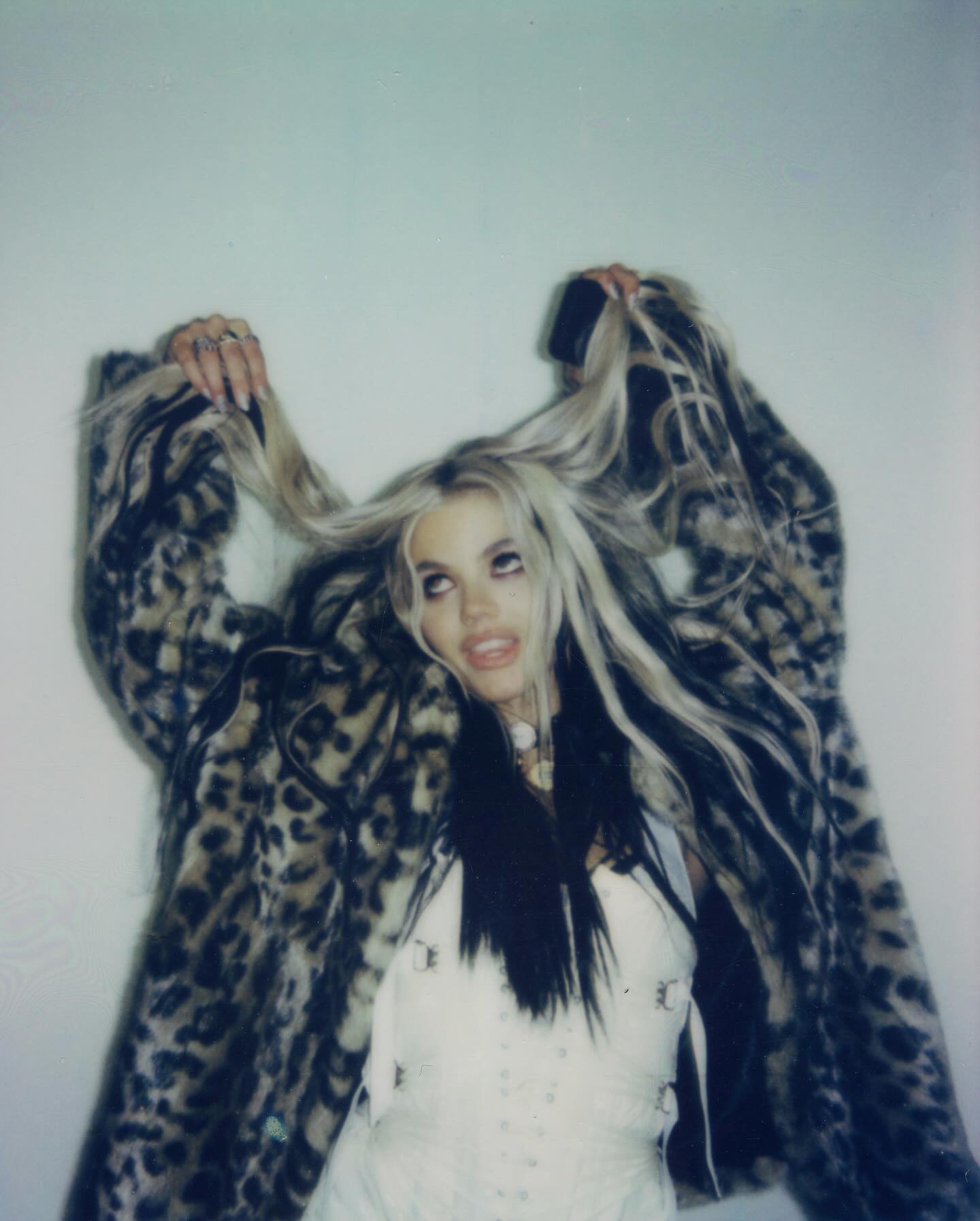 a woman with long, blonde hair with black streaks, wearing a white corset and a leopard print fur coat, is pictured in a lowangle shot. she is holding sections of her hair up with her hands, looking upwards with an open mouth. the image has a slightly faded, vintage aesthetic. she's wearing several rings.