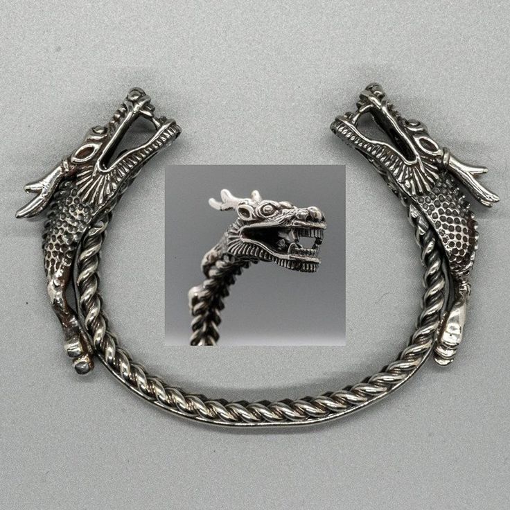 a silver dragon bracelet is featured in the image. the bracelet is shaped like a c, with a dragon head at each end. the dragons have intricate details such as scales and teeth. the band of the bracelet has a braided design. in the center of the bracelet, there is a closer shot of one of the dragon heads. the image is set against a gray background.