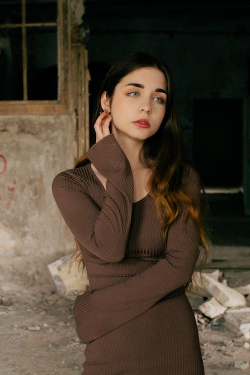 a young woman with long brown hair is captured in a thoughtful pose in an abandoned building. she's wearing a brown ribbed longsleeved dress and has one hand gently touching her face. the setting enhances the mood of the image, creating a vintage or retro feel. the lighting is dramatic, highlighting the woman's features and the texture of the dress against the dilapidated background.