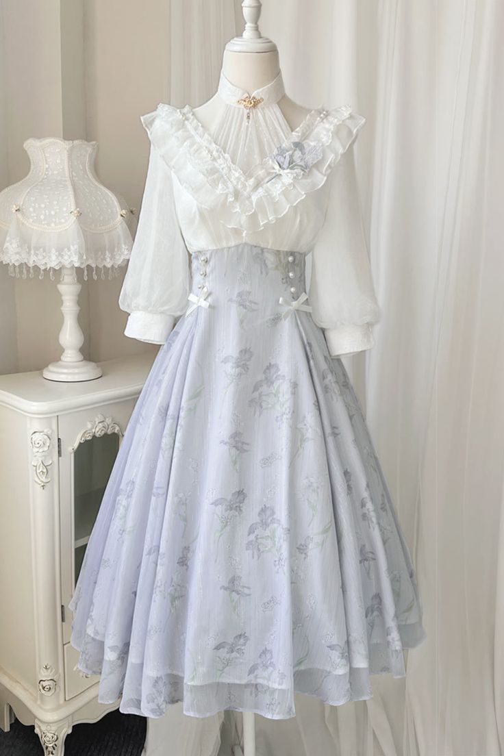 a fulllength view of a white and purple floral lolita dress displayed on a mannequin. the dress features a lace bodice with a high collar and ruffled sleeves, and a flowing skirt with a floral print. the background is a white room with a white cabinet to the left.