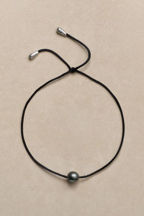 a simple and elegant necklace with a single black pearl pendant on a black string. the necklace is tied with silver ends and hangs against a light beige background.