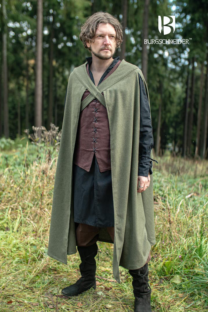 a man stands in a forest, wearing medievalstyle clothing. he is wearing a dark green cape, a brown tunic with laced detailing, a black skirt, and black boots. the burgschneider brand logo is visible in the upper right corner of the image.