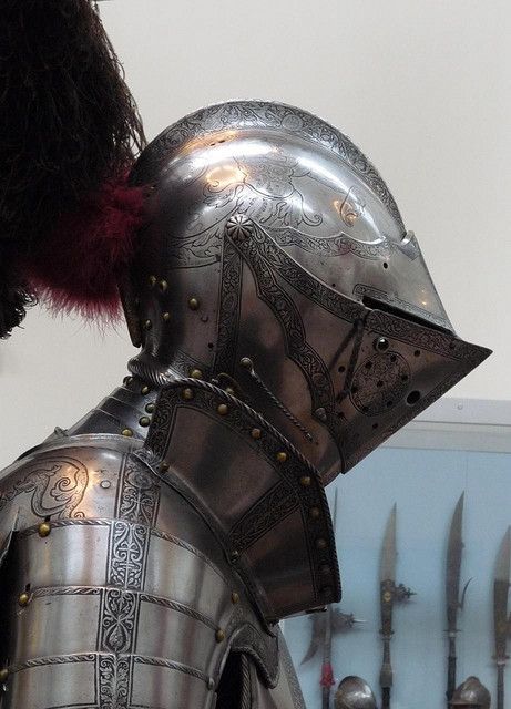 a closeup view of an ornate suit of armor, focusing on the helmet and upper torso. the armor is made of polished steel with intricate etchings. the background is a museum display case with swords and other weaponry visible, suggesting a historical context.
