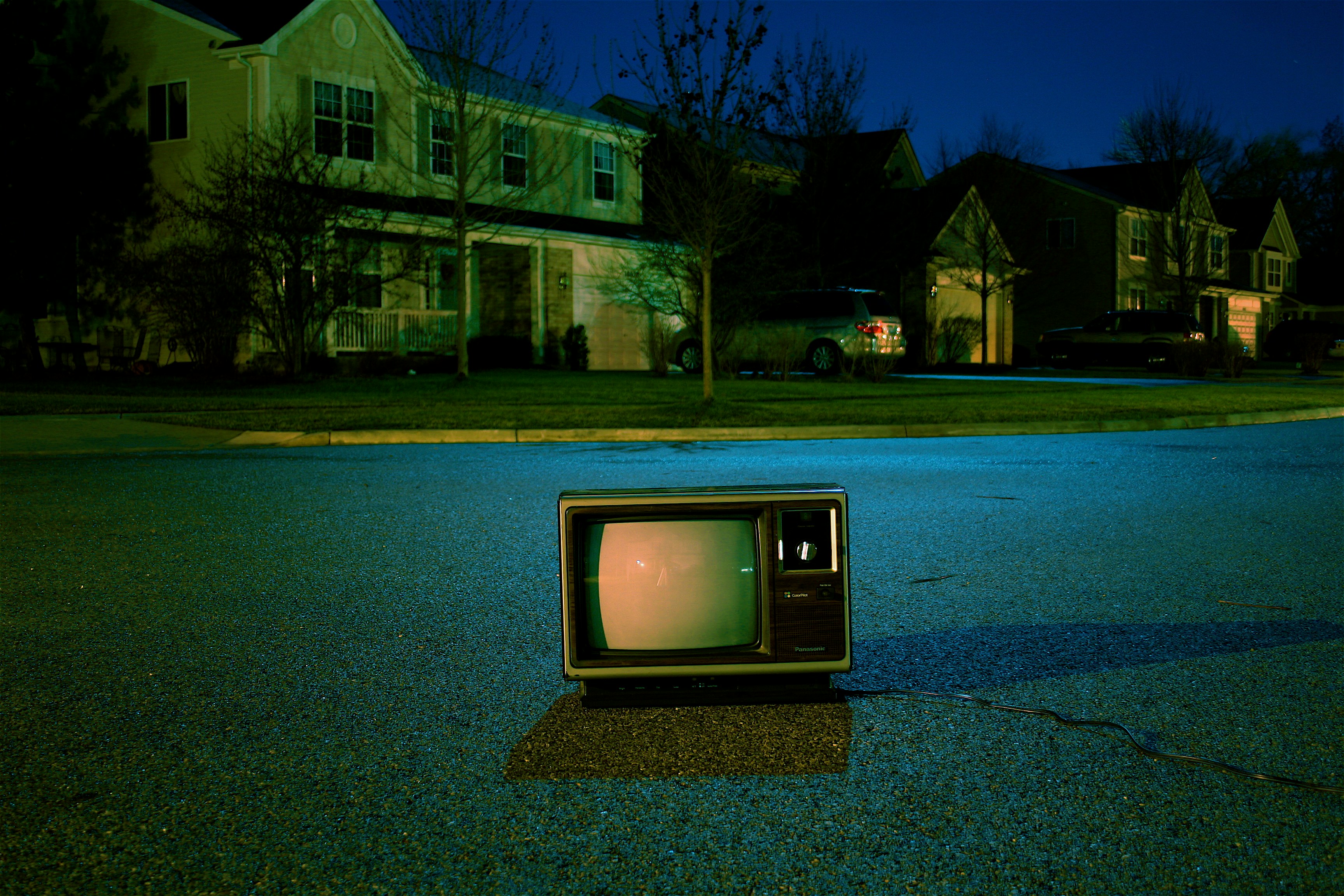 an old television sits abandoned on a suburban street at night. the television is on, emitting a faint glow, but the screen is distorted and unclear. the houses in the background are dark, with only a few lights visible. the overall atmosphere is eerie and mysterious.