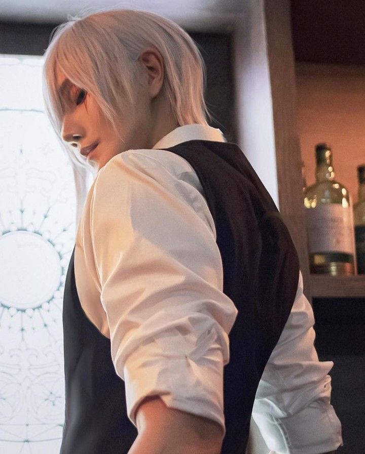 a young man in an anime cosplay is looking down. he has white hair, a white shirt, and a black vest. the background is a blurry image of a bar with liquor bottles on the shelves.