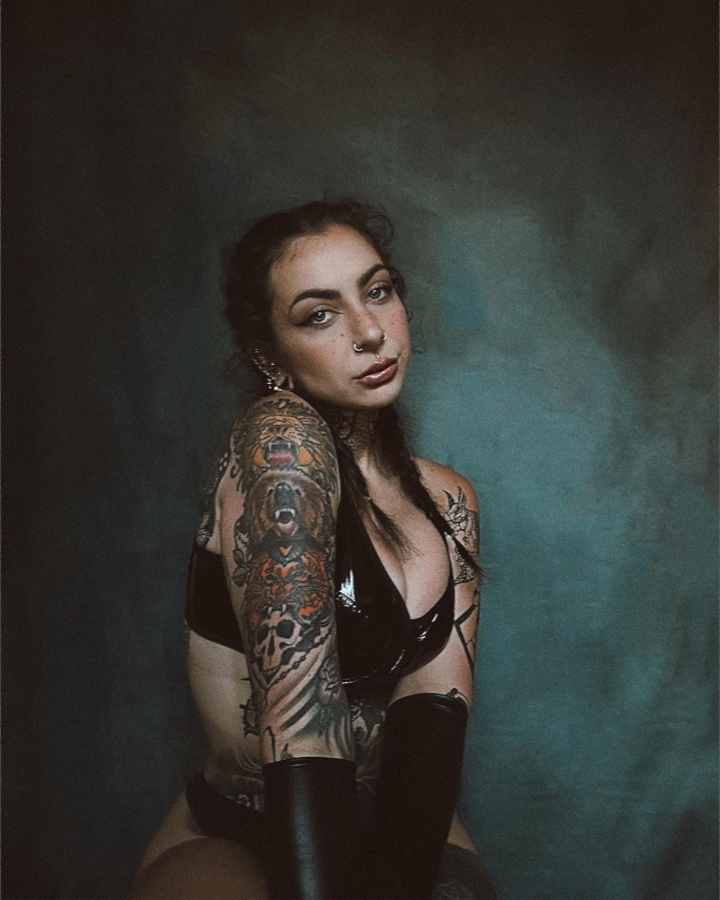 the image is a portrait of a young woman with extensive tattoos on her arms. her tattoos are detailed and feature bears, skulls, and floral elements in a style that suggests a blend of traditional and neotraditional tattooing. she's wearing a black leatherlike bra and long black gloves, contributing to a dark and edgy aesthetic. her expression is serious and intense, her gaze directly meeting the viewer's.  the background is a dark, moody teal, which enhances the overall dramatic tone of the photograph. the lighting is carefully controlled, highlighting her features and tattoos.  the overall style suggests a fashion or editorial portrait.