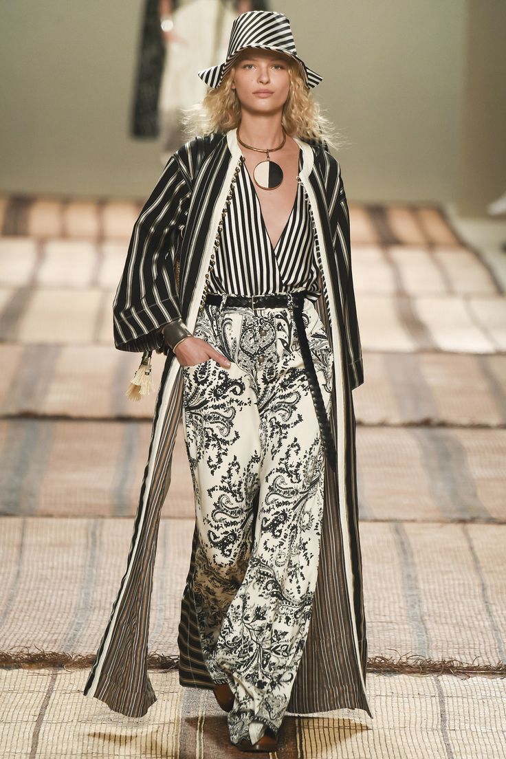 a fashion model walks down a runway wearing a stylish black and white outfit. the outfit includes a long striped coat, wideleg paisley pants, a striped top, and a black and white striped hat. she is also wearing a unique circular necklace. the overall style is bohochic and elegant. the background shows a textured runway.
