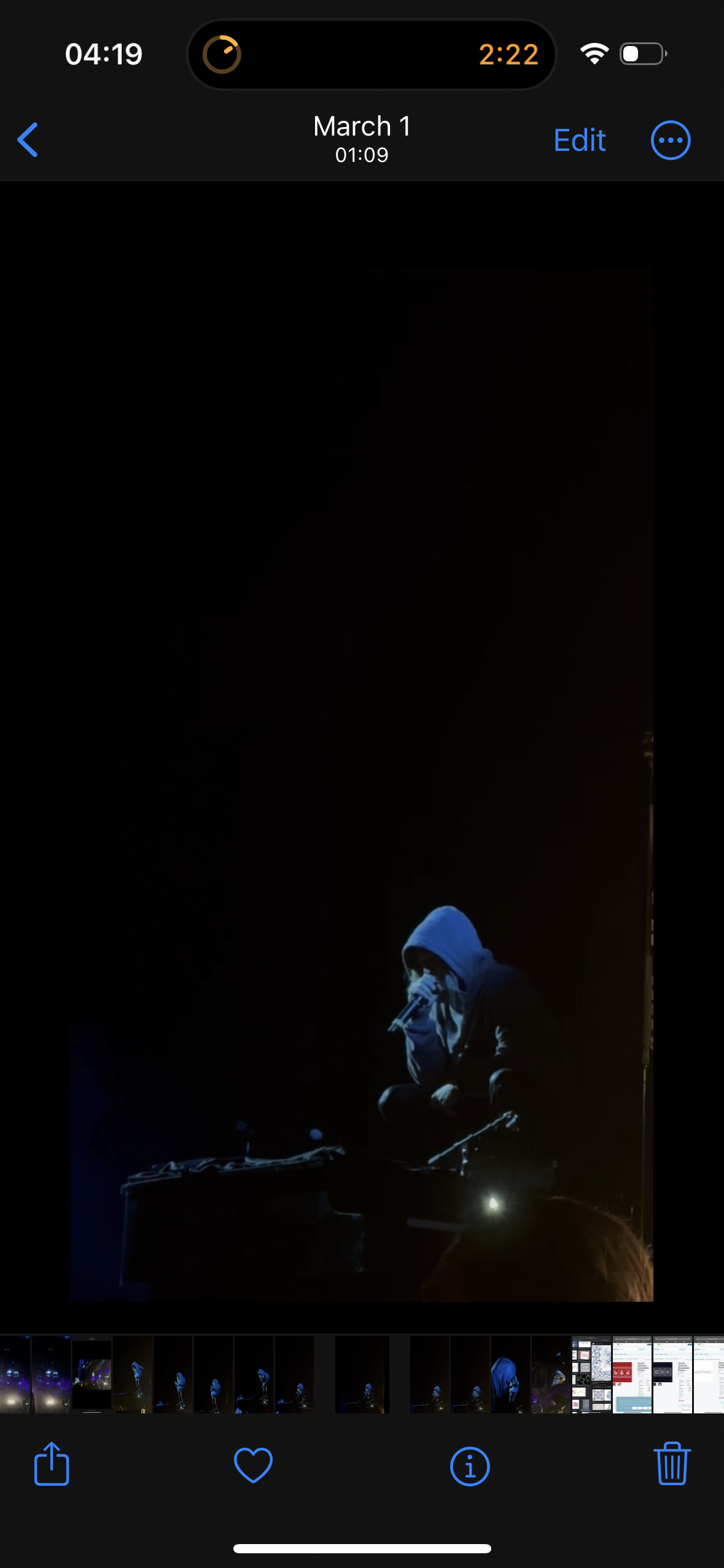 a person in a blue hoodie and dark clothing is on a stage performing in front of a dark crowd. they are singing into a microphone and the spotlight shines on them from the right. the stage is a dark wooden platform with a dark backdrop behind the performer. the image is very dark and the performer's face is not visible. the image is taken from a phone and there is a small row of thumbnails across the bottom.
