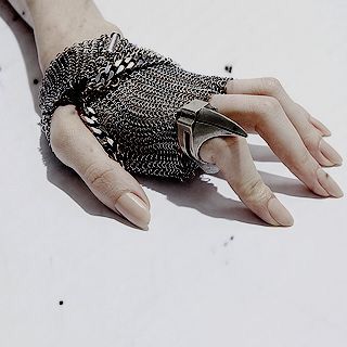 a hand wearing a silver chainmail glove with a pointy silver ring with a claw design. the ring is on the middle finger. the hand is laying flat on a white surface, with the fingers spread apart. the fingernails are painted a nude color. the hand and ring are in focus, while the background is blurry. the overall aesthetic is chic and elegant.