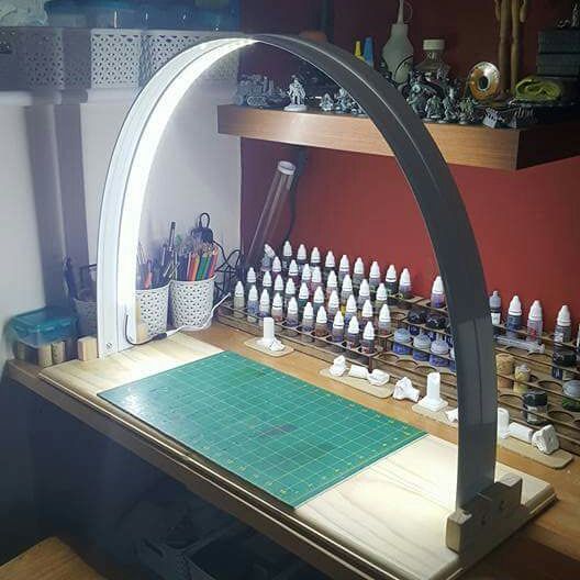 the image shows a wellorganized crafting desk with various hobby supplies. an archshaped led lamp illuminates the workspace, which includes a green cutting mat, multiple paint bottles arranged in a wooden organizer, miniature figures on a shelf above, and containers holding pencils and paintbrushes. the desk appears to be made of wood and is used for miniature painting or a similar craft.