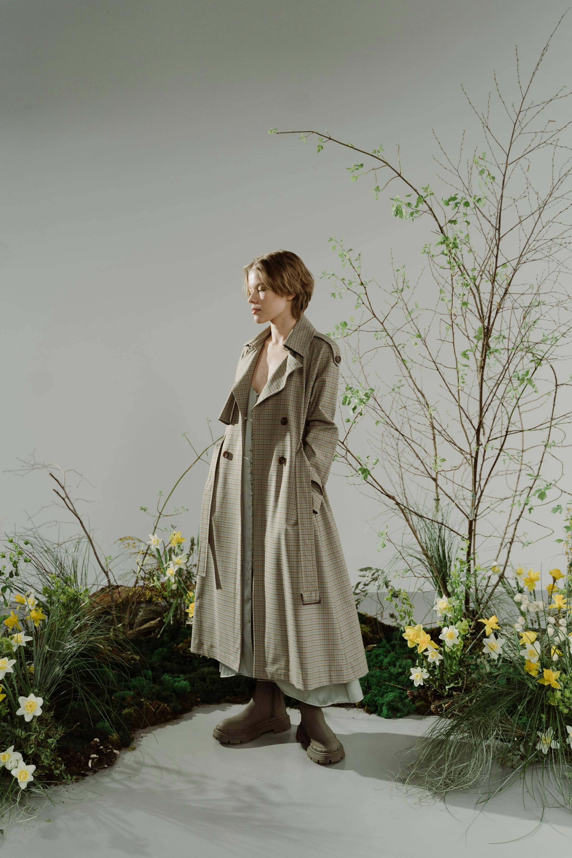 a fullbody shot of a woman in a stylish light beige and grey checked trench coat, standing amidst a spring garden setting.  she's wearing beige boots and is looking down. the background features blossoming daffodils and other greenery, creating a serene and fashionable atmosphere. the overall lighting and composition suggest a professional photoshoot.