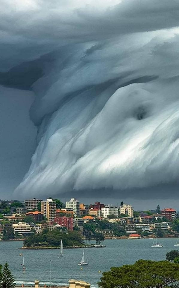 a dramatic shelf cloud hangs low over the city,  making it appear as if it is about to engulf the city below. the city is situated on the coast, and there are sailboats in the water.