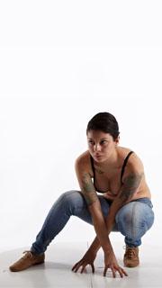 a woman with dark hair and tattoos is crouching on the floor against a white studio backdrop. she is wearing jeans and a black bra, and her gaze is directed offcamera. the soft lighting emphasizes her form and the details of her tattoos.  the low camera angle creates a sense of strength and confidence in the pose.