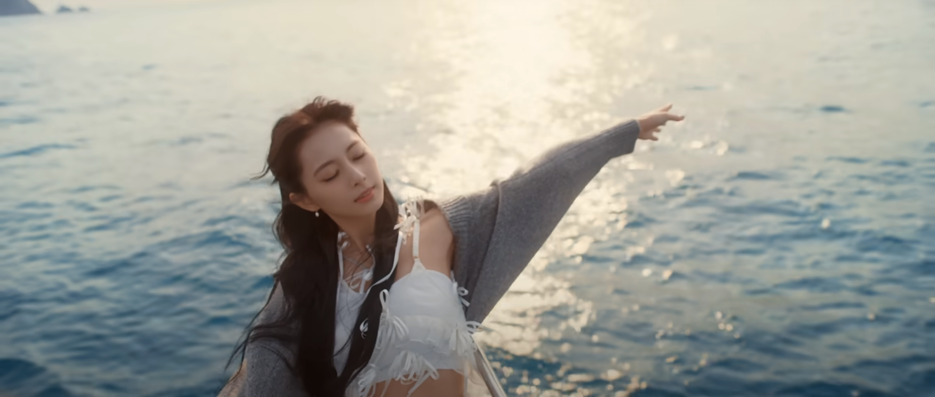 a young woman with long black hair stands on a boat and extends her arm out towards the ocean. she is wearing a white top, a gray sweater, and her eyes are closed. the image is backlit by the setting sun, which casts a warm glow on the water.