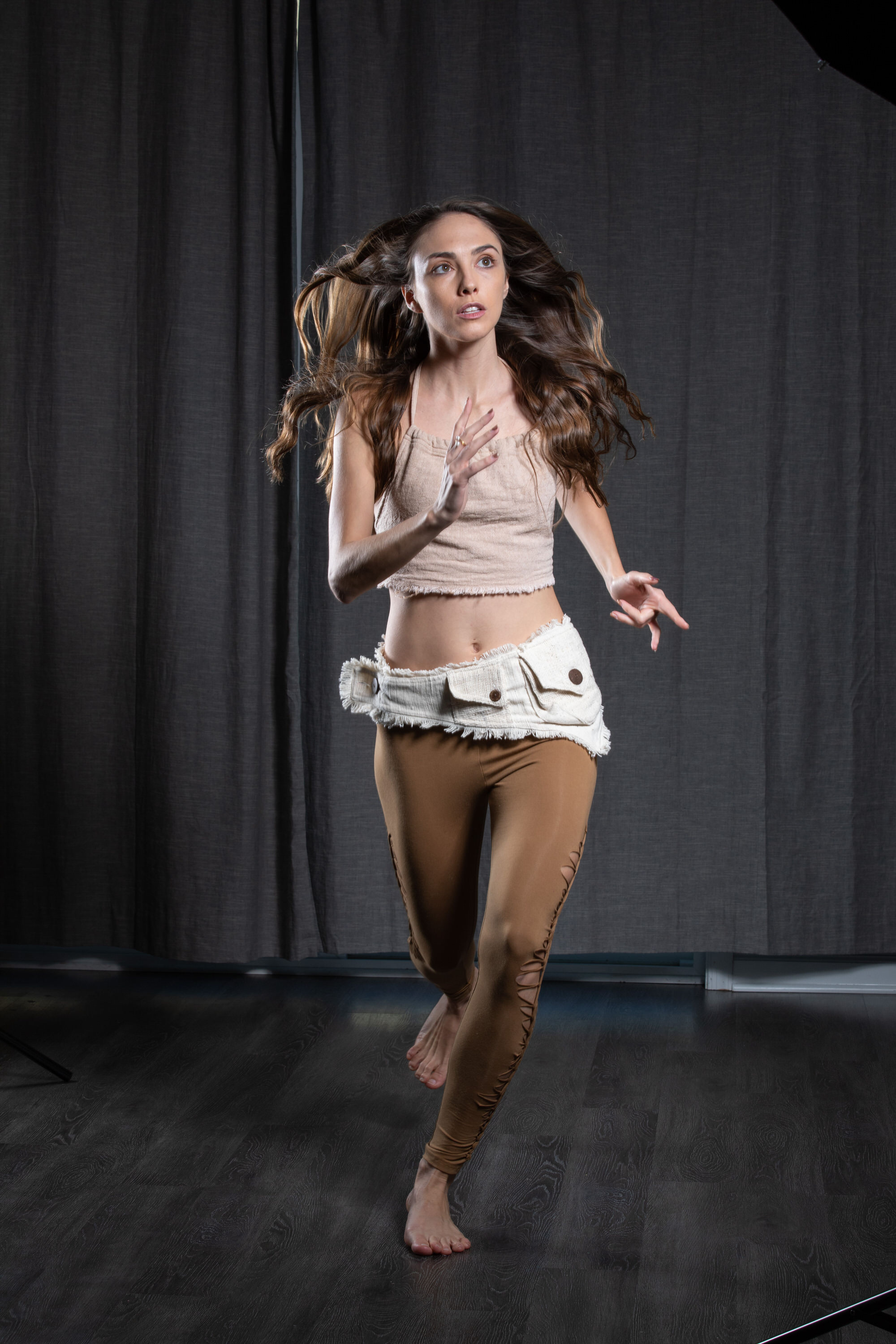 a young woman with long brown hair is captured midrun against a dark studio backdrop. her expression suggests fear or urgency, with wide eyes and an open mouth. she is barefoot and dressed in a bohemian style outfit featuring a beige crop top and brown leggings with a white fabric belt. the image is welllit with studio lighting, providing a clear view of the subject and her dynamic movement.