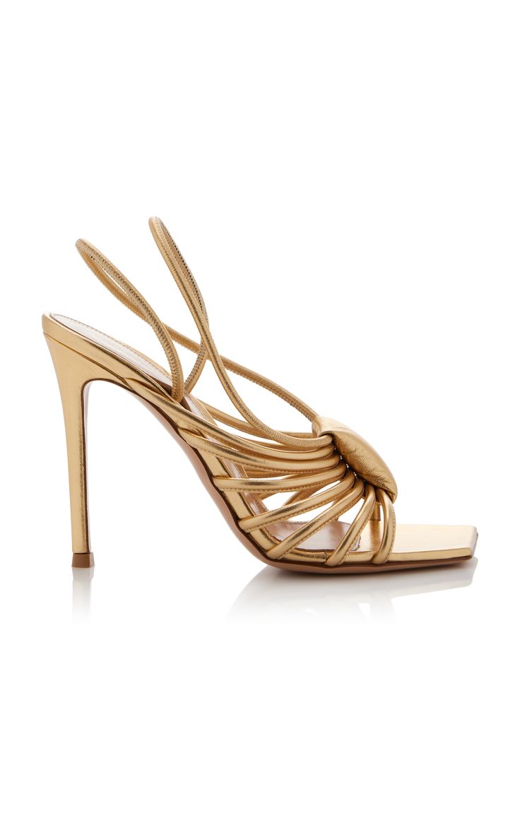 a pair of elegant gold metallic leather slingback sandals are showcased. the shoes feature a high stiletto heel and square toe. the straps are intricately knotted and crisscross the foot, adding a unique and stylish touch. the overall aesthetic is sophisticated and glamorous, perfect for special occasions or formal events. the sandals are presented on a white background, highlighting their design and luxurious details.