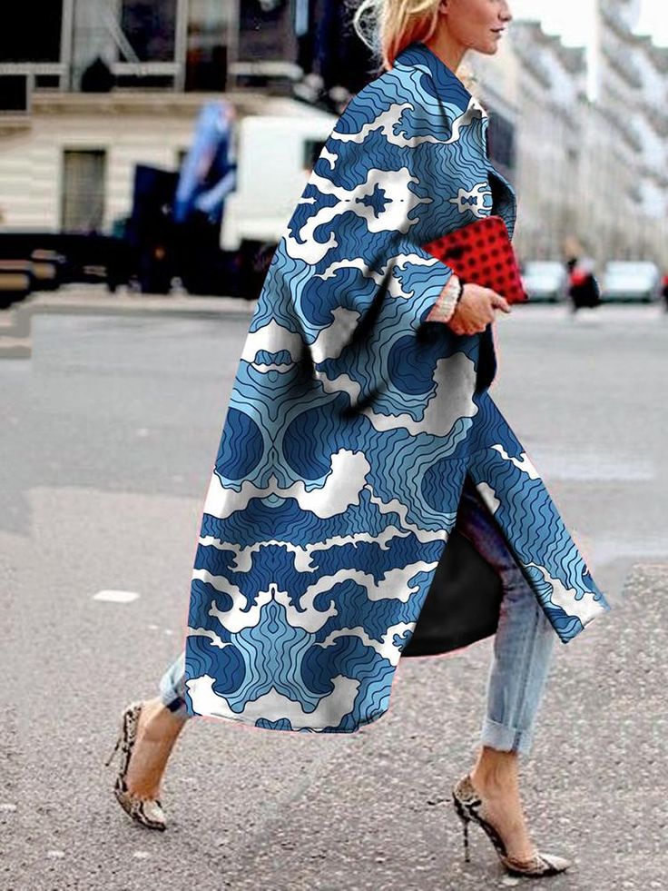 a fulllength shot of a stylish woman wearing a long, blue coat with a wave print. she is walking down a city street, carrying a red handbag. she is wearing jeans and high heels. the coat is loosefitting and has a relaxed silhouette. the woman's blonde hair is partially visible. the background is slightly blurred but shows a city street scene.