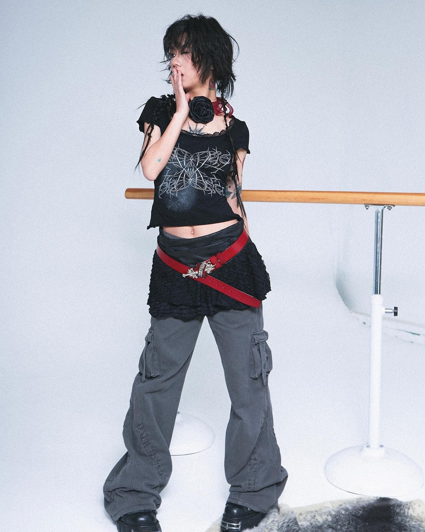 a young woman with short black hair is standing in a studio against a white background. she is wearing a black crop top with a graphic design, a red belt, grey cargo pants, and black shoes. she is also wearing a black rose necklace and has tattoos on her arms. she is posing with one hand covering her mouth.  a wooden ballet bar is visible in the background.  a furry rug is partially visible at her feet.  the overall style is grunge or alternative.