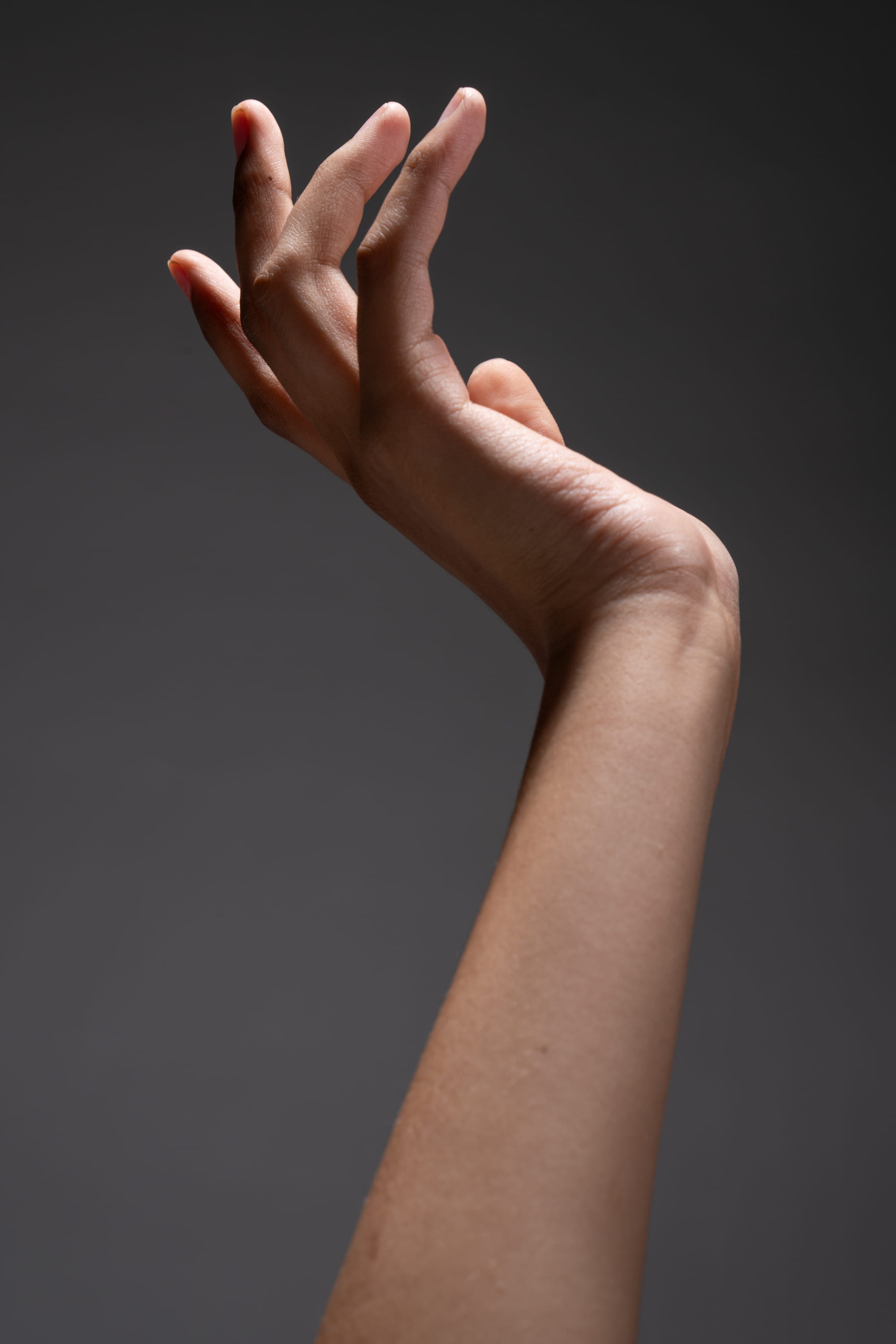 the image showcases a single female hand and arm gracefully extended upwards against a neutral gray backdrop. the hand is delicately posed with fingers slightly spread, emphasizing its form and texture. a spotlight illuminates the hand and arm, creating a dramatic contrast of light and shadow.