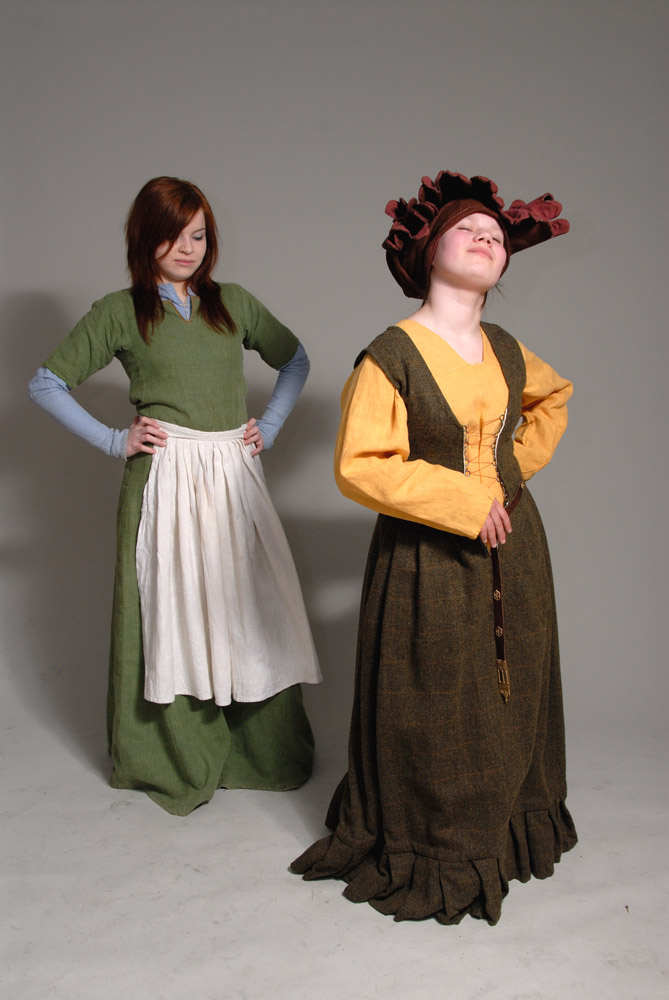 two women are wearing historical clothing. the woman on the left is wearing a long green dress with a white apron and blue longsleeved shirt underneath. the woman on the right is wearing a long yellow dress with a brown bodice, a brown belt, and a brown hat with ruffles. they are both standing in front of a gray background.
