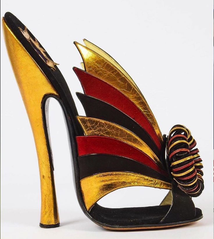 a single highheeled shoe is displayed against a plain white background. the shoe is a striking combination of gold, red, and black materials, featuring a tall, slender gold heel and a toe portion covered in black fabric. the design is particularly eyecatching due to the decorative "wing" like details, which are layers of gold, red, and black, fanning out from the middle of the shoe. a bow or twisted ornament, matching the shoe's colors, embellishes the front of the shoe near the toe. the overall impression is one of opulence and artistry.