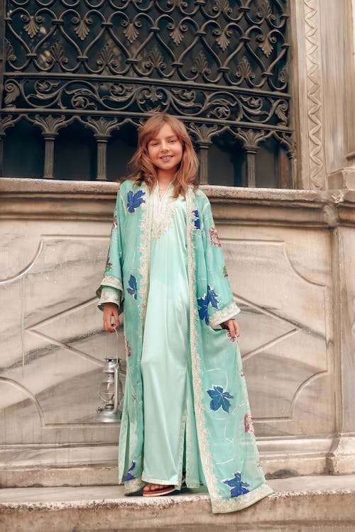 a young girl, smiling, stands on a marble step in front of an ornate stone building. she wears a beautiful mint green robe or kimono with a floral pattern and embroidered details.  she is holding an antique lantern. the building features an intricately designed wroughtiron window. the overall aesthetic suggests a historical or cultural setting, possibly in the middle east. the girl's clothing is elegant and traditional in style.