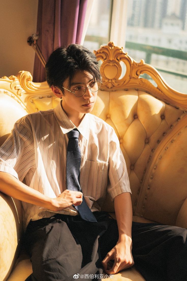 a young man is sitting on a luxurious goldcolored armchair in a sundrenched room. he's wearing a white shortsleeved shirt with thin black stripes, black pants, and a dark blue tie, which he is adjusting with his hand. he is also wearing stylish glasses and looking thoughtfully into the distance. the sunlight creates a warm and inviting atmosphere in the image.