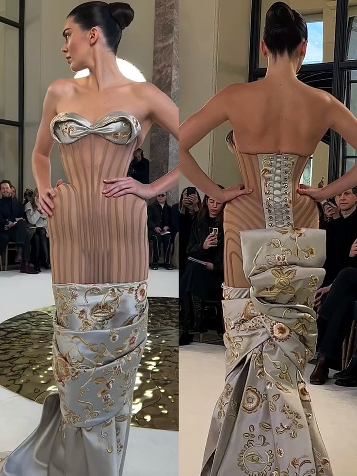 the image showcases model kendall jenner in a highfashion dress during a fashion show, with front and back views displayed side by side. the dress features a strapless corset top, a structured bodice, and a detailed floralembroidered skirt that wraps around the body. jenner has her hair styled in a neat high bun. the audience and onlookers can be seen in the background, observing the runway presentation. the dress consists of a tan corset with a silver bustier piece and silver lacing in the back, adorned with tan fabric with vertical stripes.
